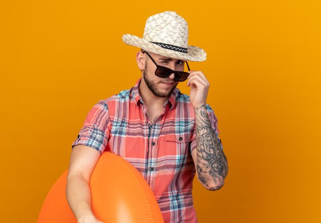 confident young traveler man with straw beach hat in sun glasses blinks his eye and holds swim ring isolated on orange wall with copy space