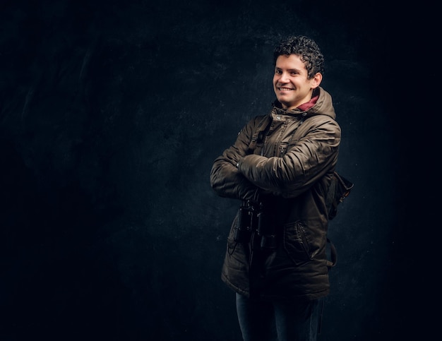 Free photo confident young traveller in hiking gear with binoculars stands with crossed hands, smiling and looking at a camera in the studio against a dark wall