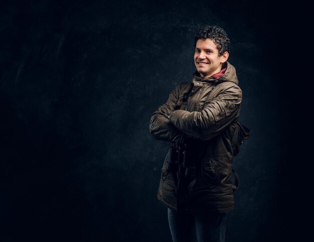 Confident young traveller in hiking gear with binoculars stands with crossed hands, smiling and looking at a camera in the studio against a dark wall