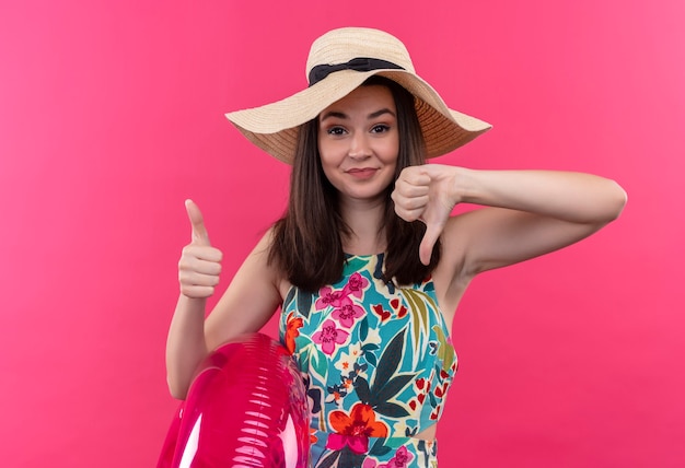 Confident young woman wearing hat holding swim ring and showing thumbs up and down on isolated pink wall