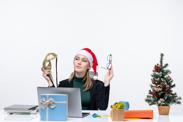 Confident young woman with santa claus hat eyeglasses and mask sitting at a table with a Xsmas tree and a gift on it in the office on white background