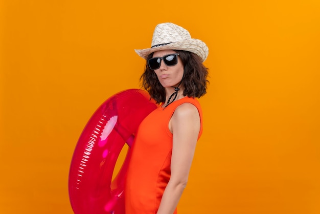 A confident young woman with short hair in an orange shirt wearing sun hat and sunglasses holding pink inflatable ring 