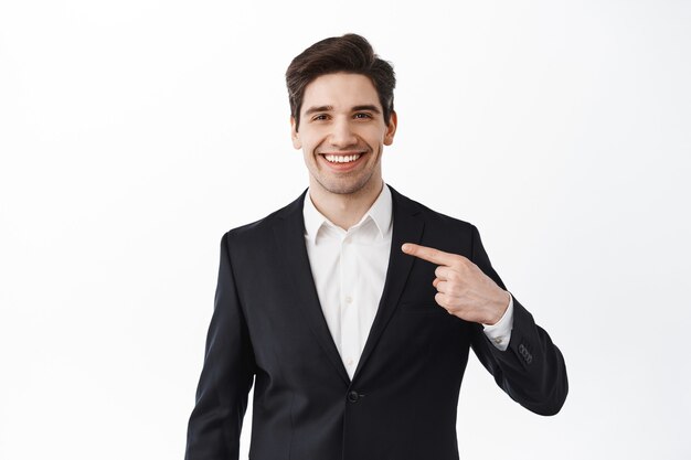 Confreal estate agent points at himself with pleased, determined smile, self-promoting, choose me gesture, stands against white wall