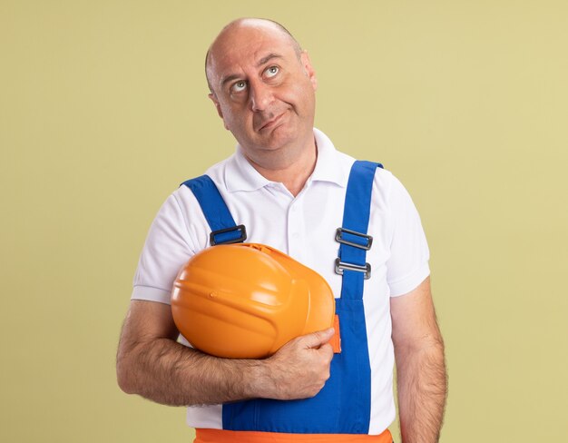 Confused adult builder man in uniform holds safety helmet isolated on olive green wall
