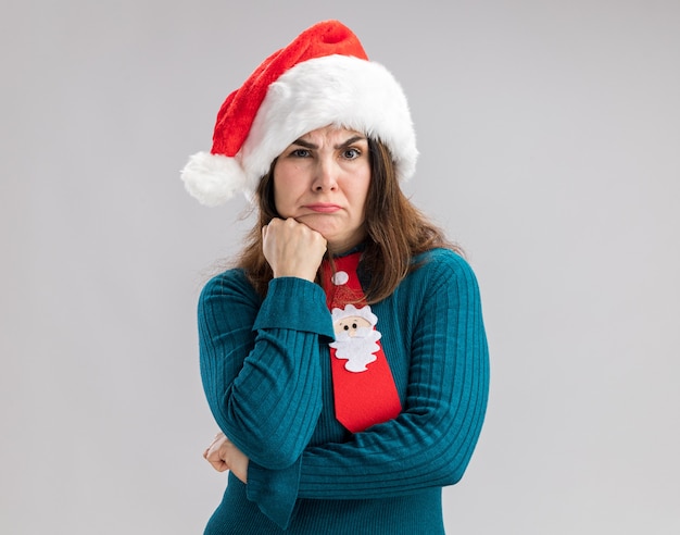 Confused adult caucasian woman with santa hat and santa tie puts fist on chin  isolated on white wall with copy space