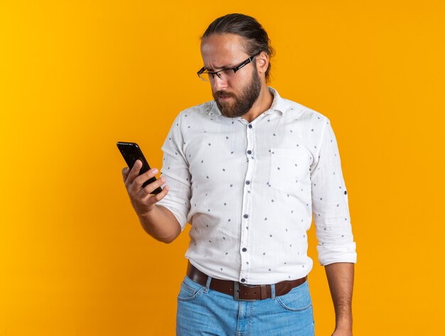 Confused adult handsome man wearing glasses holding and looking at mobile phone isolated on orange wall