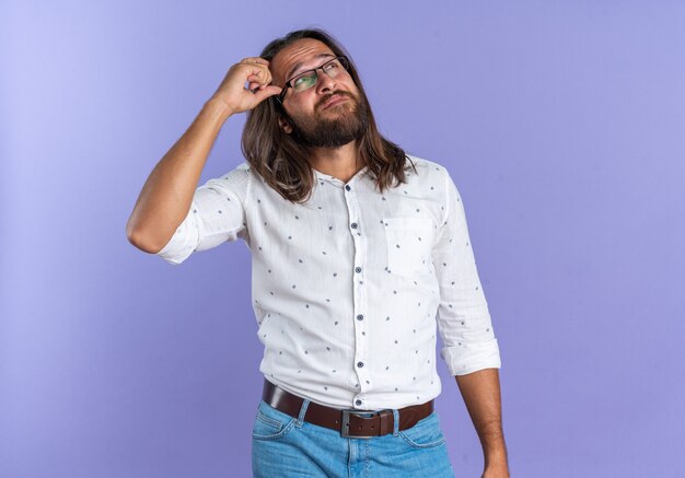 Confused adult handsome man wearing glasses keeping hand on head looking up isolated on purple wall with copy space