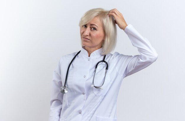 Confused adult slavic female doctor in medical robe with stethoscope putting hand on head isolated on white background with copy space