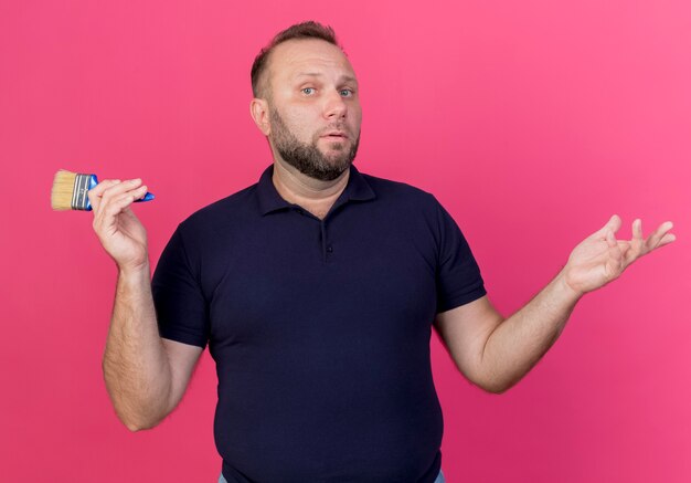 Confused adult slavic man holding paint brush and showing empty hands isolated