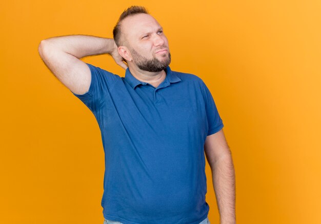Смущенный взрослый славянский мужчина смотрит в сторону, держа руку за шею, изолированную на оранжевой стене с копией пространства