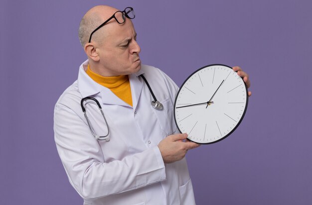 Confused adult slavic man with optical glasses in doctor uniform with stethoscope holding and looking at clock