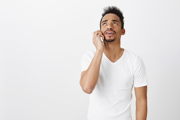 Confused african american guy in white t-shirt talking on mobile phone, looking up puzzled or uncertain