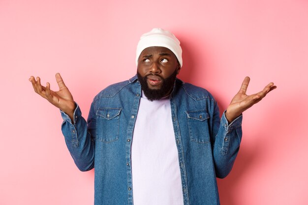 Confused african-american man in beanie looking at upper left corner doubtful, shrugging uncertain, standing over pink background