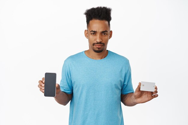 Confused african american man furrow eyebrows, showing credit card with mobile phone empty screen, looking doubtful or disappointed, standing against white background