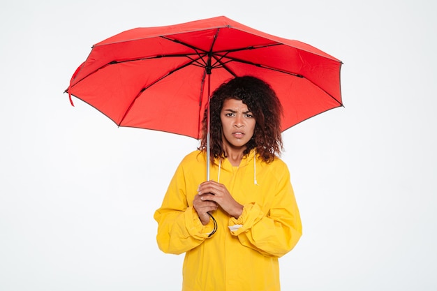 Confused african woman in raincoat hiding under umbrella