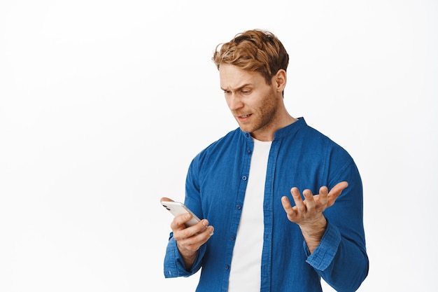 Бесплатное фото Смущенный и раздраженный молодой человек смотрит в смартфон, жалуется на приложение, читает надоедливое нелепое сообщение, поднимает раздраженную руку, стоит над белой стеной