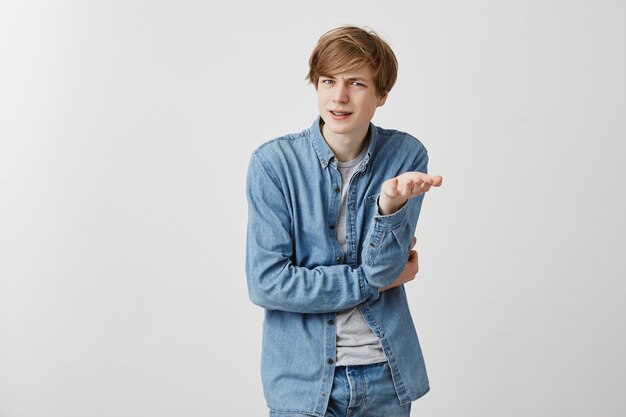 Confused angry fair-haired young male student gestures in bewilderment, expresses negative emotions as has conversation with girlfriend who critises him. Good-looking caucasian guy in confusion