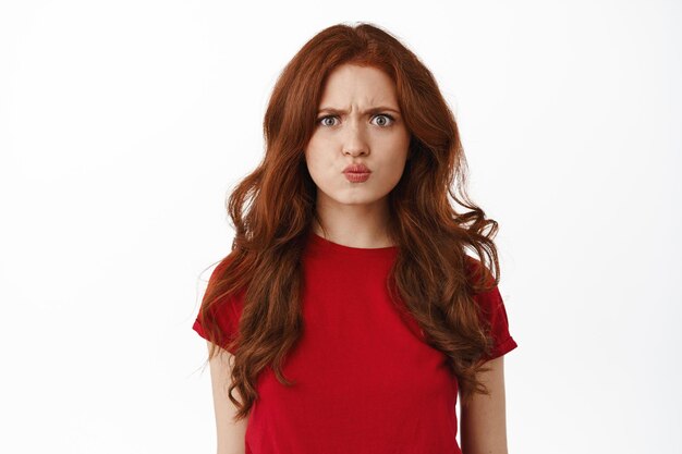 Confused and angry redhead girl pouting, frowning and grimacing, looking upset, sulking offended, being insulted or displeased, standing against white background.