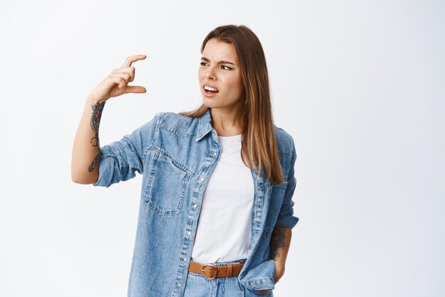 Confused and annoyed young woman frowning staring at small size gesture disappointed complaining on little thing standing against white background