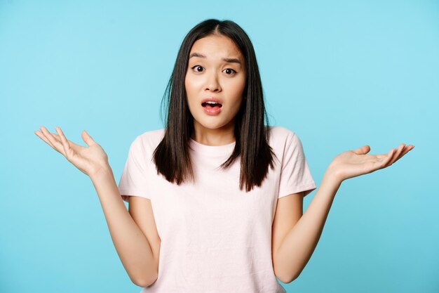 混乱したアジアの女の子が空の手を横に広げて肩をすくめ、Tシャツの上に無知に立っています...