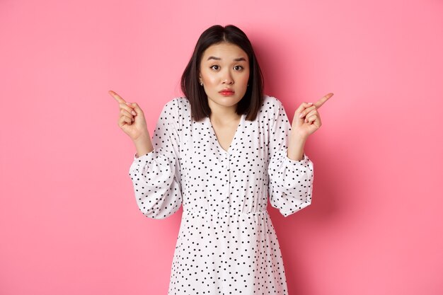혼란스러운 아시아 여성은 주저하고 어깨를 으쓱하고 손을 양쪽으로 가리키며 우유부단한 표정을 짓고 선택에 도움이 필요하며 분홍색 배경 위에 서 있습니다.