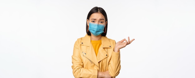 白いスタジオの背景の上に立っている医療用フェイスマスクを身に着けている無知な困惑を探している医療用フェイスマスクの混乱したアジアの女性