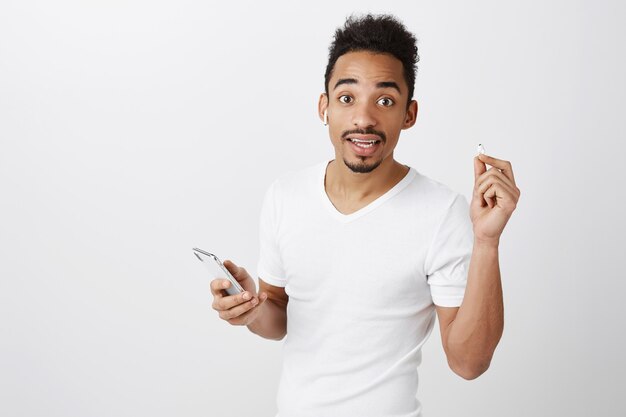 Confused attractive african american guy take-off earphone to listen, holding mobile phone, stop listening music