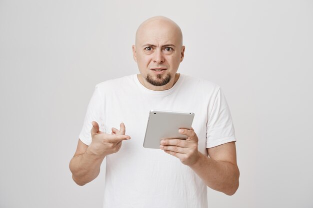 Смущенный лысый взрослый мужчина с недоумением смотрит на цифровой планшет