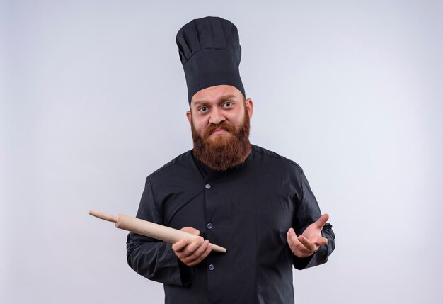 A confused bearded chef man in black uniform holding rolling pin while looking at camera on a white wall