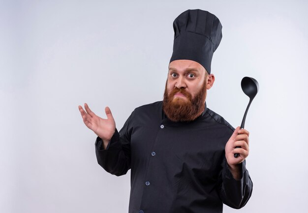 A confused bearded chef man in black uniform raising ladle on his hand on a white wall