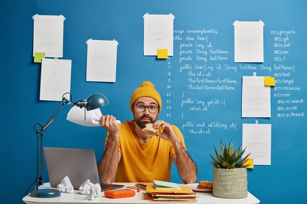 Confused bearded freelance worker develops startup project, has lunch, eats delicious snack, holds paper document in hand, poses at desktop.