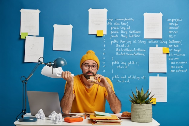 Бесплатное фото Смущенный бородатый внештатный работник разрабатывает стартап-проект, обедает, ест вкусную закуску, держит в руке бумажный документ, позирует за рабочим столом.