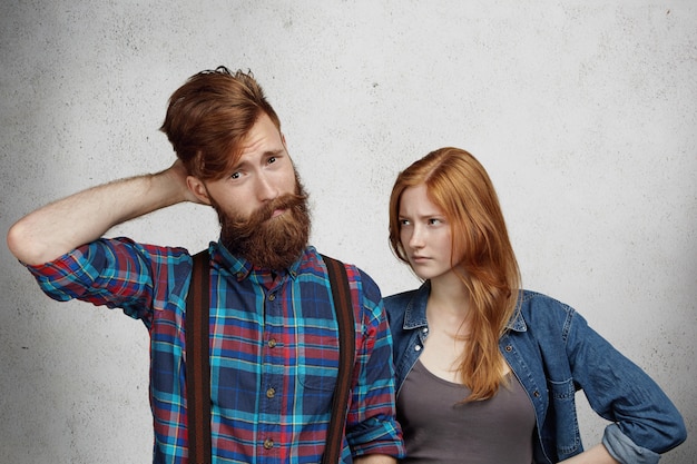 Free photo confused bearded man feeling guilty, scratching his head in confusion while his redhead girlfriend or wife standing next to him, looking with angry disappointed expression, holding hands on her waist