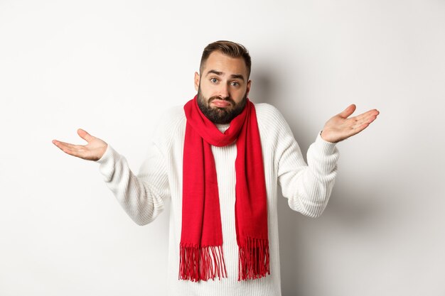 Confused bearded man shrugging, raising hands up and looking clueless, dont know anything, standing in sweater and christmas scarf, white background