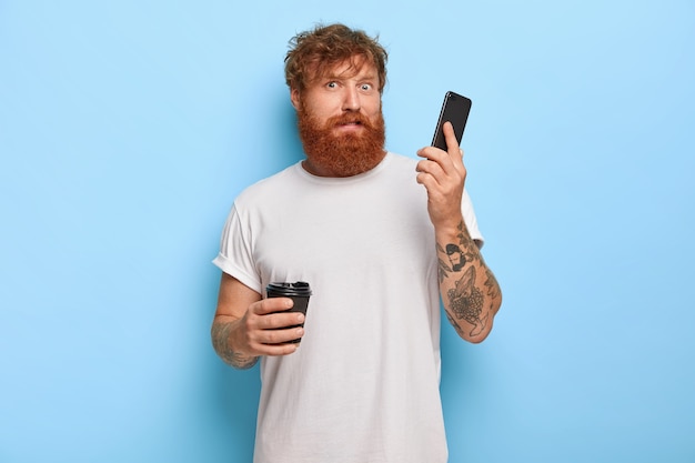 Confused bearded redhaired guy holds mobile phone, gets call from unknown person, hears terrible loud scream via cellular, drinks takeaway coffee, wears white casual t shirt