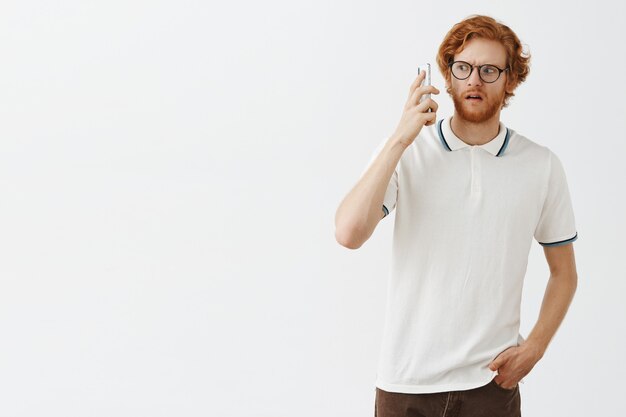 Confused bearded redhead guy posing against the white wall with glasses