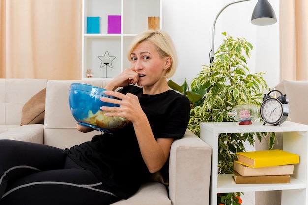 Confused beautiful blonde russian woman lying on armchair biting fist and holding bowl of chips looking at camera inside the living room