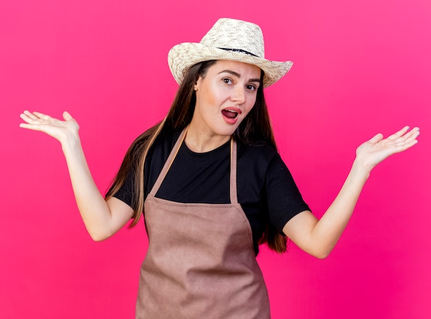 Confused beautiful gardener girl in uniform wearing gardening hat spreading hands isolated on pink background
