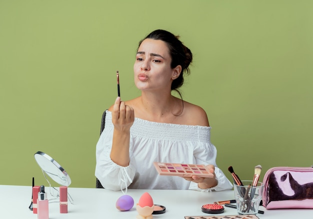 Confused beautiful girl sits with closed eyes at table with makeup tools holding eyeshadow palette and makeup brush isolated on green wall