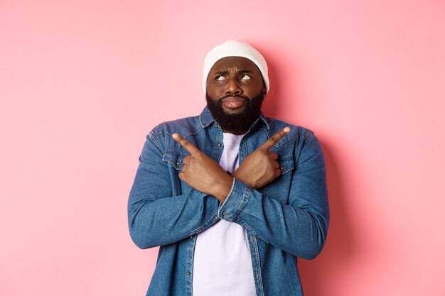 Confused Black man with beard, making choice, pointing fingers sideways and looking puzzled, standing over pink background.