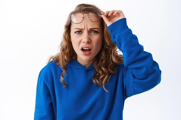 Confused blond girl take off glasses look with puzzled and displeased face cringe from something strange and awful standing in blue hoodie against white background