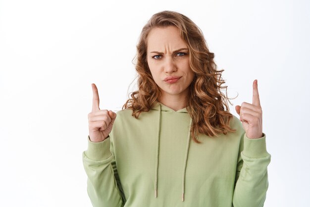 Confused blond woman frowning, judging bad promo, pointing fingers up and look with skeptical disappointed face expression, standing over white wall