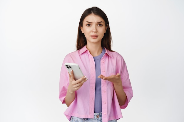 Confused brunette woman pointing at her phone with puzzled and concerned face expression white background