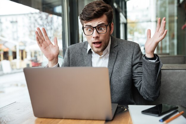 Confused businessman in eyeglasses sitting by the table in cafe and looking at laptop computer