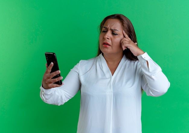 Confused casual caucasian middle-aged woman holding and looking at phone putting finger on forehead 