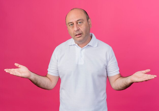 Confused casual mature businessman looking at camera and showing empty hands isolated on pink background