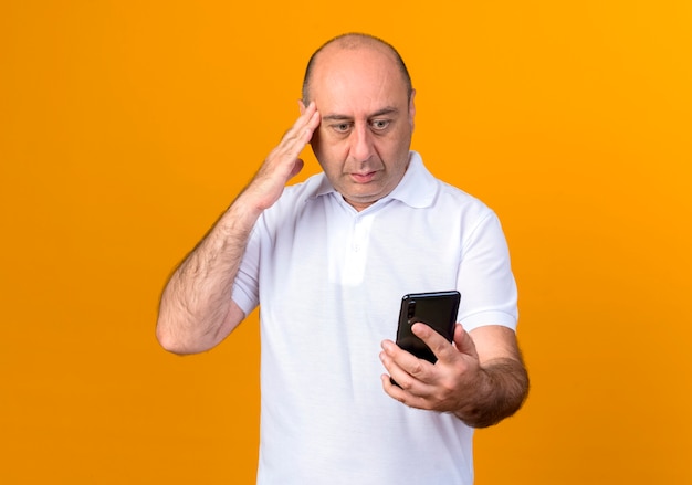 Confused casual mature man holding and looking at phone putting hand on temple isolated on yellow backgound