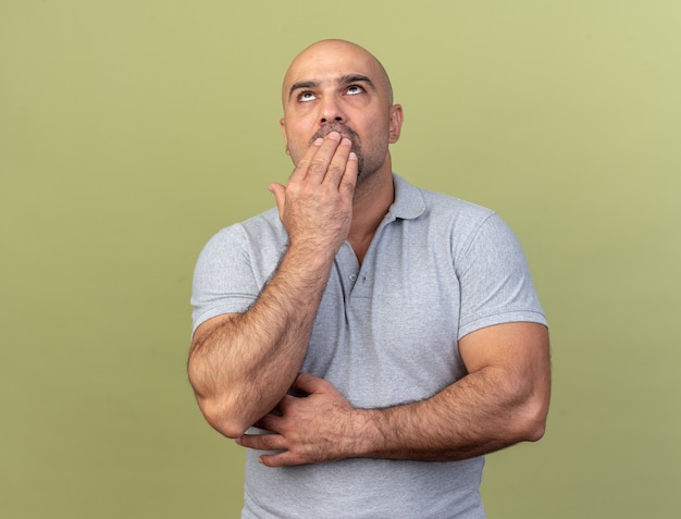 Confused casual middle-aged man keeping hand on mouth looking up isolated on olive green wall