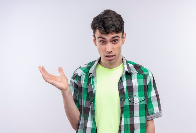 Confused caucasian young man wearing green shirt held out at side hand on isolated white wall