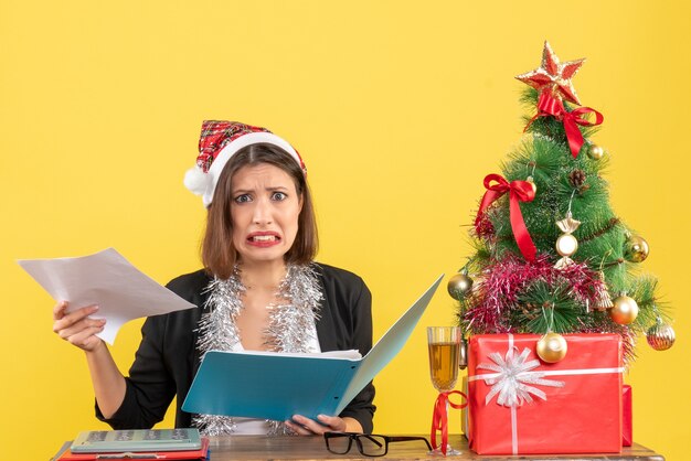 Confused charming lady in suit with santa claus hat and new year decorations holding document in the office on yellow isolated 